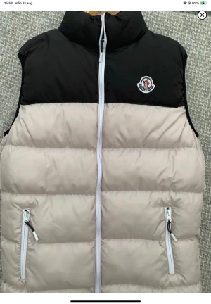 Beige och svart dunväst från Moncler - Snygg dunväst från Moncler i beige och svart med hög krage och två dragkedjeförsedda fickor. Västen har en vit dragkedja framtill och Moncler-logga på bröstet. Perfekt för lager på lager och riktigt trendig look.