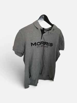 Grå piké från Morris - Säljer en grå pikétröja från Morris med svart broderad logga och texten 'EST 1968' på bröstet. Klassisk krage och korta ärmar, perfekt till en avslappnad stil. Tillverkad i mjuk bomull för skön känsla.