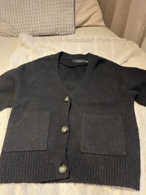 Svart stickad kofta med fickor - Mysig svart stickad kofta med v-ringning och tre stora knappar framtill. Koftan har två rejäla fickor och ribbade muddar vid ärmslut och nederkant. Perfekt att slänga över en t-shirt för en avslappnad look.