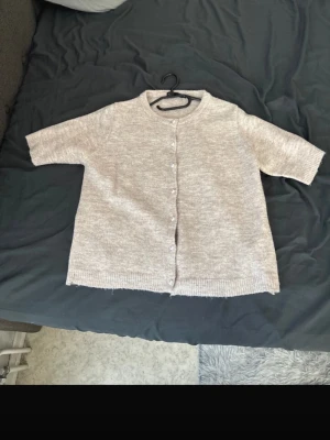 Beige kofta med kort ärm från 157 - Mysig beige kofta från 157 med korta ärmar och rund hals. Koftan har knäppning framtill med små knappar och är stickad i ett mjukt material. Perfekt att slänga över en topp för en chill look.