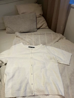 Vit kofta med pärlknappar från 157 - Kortärmad vit kofta från 157 med rund halsringning och pärlknappar framtill. Koftan har ribbade kanter och är gjord i ett mjukt stickat material som känns skönt mot huden. Perfekt att styla över en topp eller klänning.