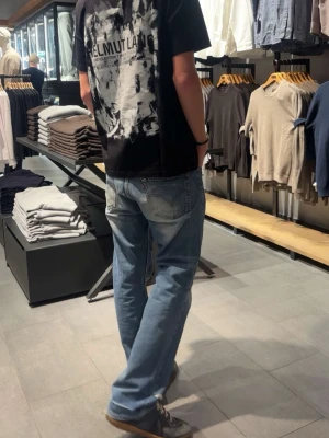 Svart Helmut Lang t-shirt med tryck - Säljer en svart t-shirt från Helmut Lang med stort vitt och grått grafiskt tryck på ryggen. T-shirten har normal passform och korta ärmar. Perfekt för dig som gillar streetwear och vill sticka ut med en unik design.