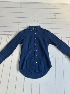 Marinblå linneskjorta från Polo Ralph Lauren - Säljer en marinblå skjorta från Polo Ralph Lauren i linne. Skjortan har klassisk button-down krage, vita knappar och den ikoniska broderade loggan på bröstet. Perfekt för en clean och stilren look.