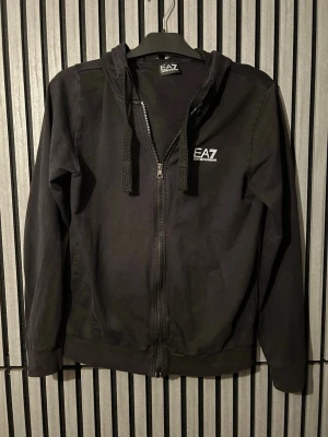 Svart hoodie från EA7 Emporio Armani - Snygg svart hoodie med dragkedja från EA7 Emporio Armani. Klassisk passform med huva och justerbara snören, använd mycket men bra skick. Pris kan diskuteras!