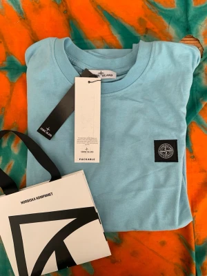 Ljusblå t-shirt från Stone Island - Säljer en ljusblå t-shirt från Stone Island med klassisk logopatch på bröstet. T-shirten har rund hals och är tillverkad i mjuk bomull. Perfekt för dig som gillar stilrena och trendiga plagg.