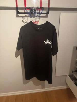 Svart Stüssy t-shirt med tryck på ryggen  - Säljer en svart t-shirt från Stüssy med deras ikoniska vita logga tryckt på bröstet. Klassisk passform och rund halsringning. Perfekt till streetwear-stilen och enkel att matcha med allt.