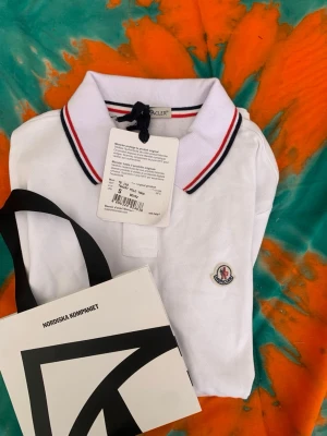 Vit pikétröja från Moncler - Klassisk vit pikétröja från Moncler med krage och knäppning framtill. Tröjan har röda och marinblå ränder på kragen och ärmen samt Monclers logga broderad på bröstet. Tillverkad i bomull för en skön och luftig känsla.