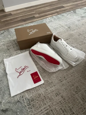 Vita sneakers med nitar från Louboutin - Snygga vita sneakers i läder från Christian Louboutin med ikoniska röda sulor och coola vita nitar på tån. Skorna har vita snören och en clean design som sticker ut. Perfekta för dig som vill ha något extra till din outfit.