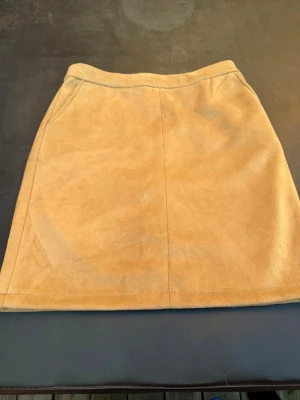 Beige mockakjol i kort modell - Snygg beige kortkjol i mockaimitation med enkel och stilren design. Kjolen har raka linjer och en diskret dragkedja bak. Perfekt att matcha med stickade tröjor eller t-shirts för en trendig look.