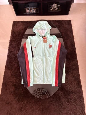 Nike Frankrike Vm kofta/jacka TRACKSUIT - Snygg mintgrön vindjacka från Nike med huva och dragkedja. Jackan har röda och mörkbruna ränder längs ärmarna samt Nike-logga och tuppemblem på bröstet. Perfekt för dig som vill sticka ut med sportig stil. Tillverkad i lätt material.