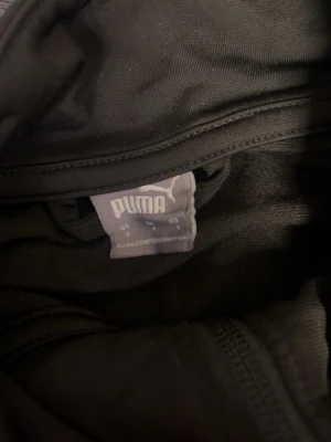 Svart träningsset från Puma. Den har lite små fläckar har bara använt byxan en gång. Kan kan användas för båda killar och tjejer! Jag kan skicka bättre bilder. - Säljer ett svart träningsset från Puma med dragkedja på jackan och diskret Puma-logga på byxan. Setet har en sportig look med elastiska muddar och är tillverkat i ett glansigt, lätt material som passar perfekt för träning eller uppvärmning.