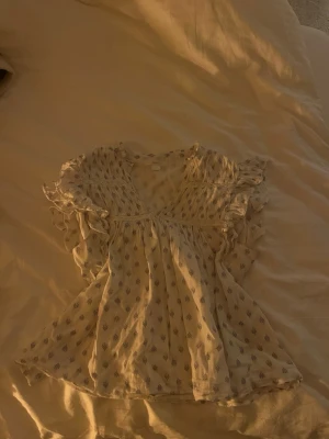 Vit blus med volang och småmönster - Söt vit blus med små blå mönster och volangdetaljer vid axlarna. Blusen har v-ringning och är luftig med korta ärmar. Perfekt för en avslappnad och trendig look🩵