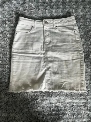 Vit jeanskjol med rå kant - Säljer en vit kort jeanskjol med rå, fransig kant nertill. Kjolen har klassiska fem fickor, bälteshällor och knappstängning framtill. Perfekt för dig som gillar en enkel men trendig look.