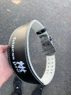 Svart träningsbälte Fitness Pro - Säljer ett svart träningsbälte från Fitness Pro med vit kontrastsöm och kraftigt spänne i metall. Bältet är gjort i skinn och har justerbara hål för perfekt passform. Perfekt för dig som vill ha extra stöd vid styrketräning.