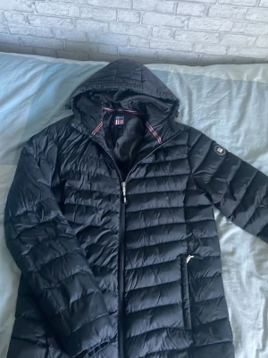 Svart dunjacka med huva - Säljer en svart lång dunjacka med huva och dragkedja framtill. Jackan har två sidofickor med dragkedja och quiltad design. Perfekt för kalla vinterdagar och riktigt skön att bära. Klassisk look som passar till det mesta.