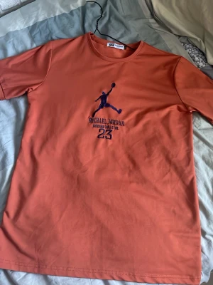 Orange Michael Jordan t-shirt - Snygg orange t-shirt med Michael Jordan-tryck och nummer 23 på bröstet. T-shirten har korta ärmar och är gjord i mjuk bomull. Perfekt för dig som gillar basket och streetwear. FUNKAR S/M