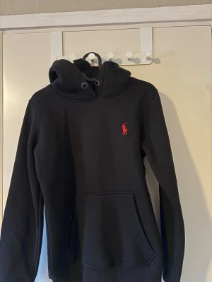 Svart hoodie från raulph lauren - Svart hoodie med huva och känguruficka framtill. Har en röd broderad logga på bröstet och breda muddar vid ärmslut och nederkant. Perfekt för en avslappnad och stilren look.
