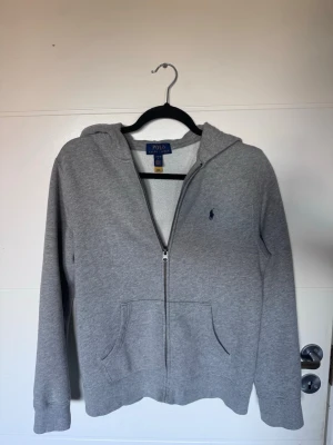 Grå zip hoodie från Polo Ralph Lauren - Säljer en grå hoodie med dragkedja från Polo Ralph Lauren. Klassisk design med huva, två fickor framtill och den ikoniska lilla loggan broderad på bröstet. Perfekt för en avslappnad och stilren look. Mjuk bomullsblandning som känns skön mot huden. Storlek XS eller 14-16 officiell storlek 