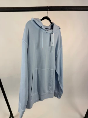 C.P company hoodie  - Snygg ljusblå hoodie med stor magficka och huva med dragsko. Ärmen har en unik detalj med en rund svart patch och liten ficka. Perfekt för dig som gillar streetwear och vill ha något bekvämt och stilrent.