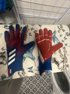 Adidas Demonskin målvaktshandskar - Säljer ett par Adidas Demonskin målvaktshandskar i blått och rött med vita detaljer. Handskarna har en tajt passform och greppvänlig insida, samt coola 3-stripes på ovansidan. Perfekta för dig som älskar fotboll och vill ha stil på planen.