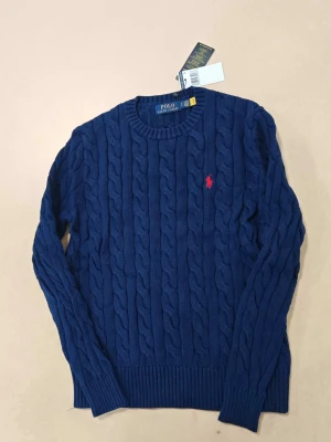 Blå kabelstickad tröja Polo Ralph Lauren - Snygg blå kabelstickad tröja från Polo Ralph Lauren med rund halsringning och röd broderad logga på bröstet. Tröjan har ribbade muddar vid ärmslut och nederkant. Perfekt för dig som vill ha en klassisk och stilren look.