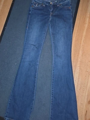 Blå bootcut jeans från True Religion - Säljer ett par blå bootcut jeans från True Religion med snygga kontrastsömmar och ikoniska fickdetaljer bak. Jeansen har låg midja, klassisk femficksdesign och coola knappar på bakfickorna. Perfekta för dig som gillar retrovibbar och streetstyle.