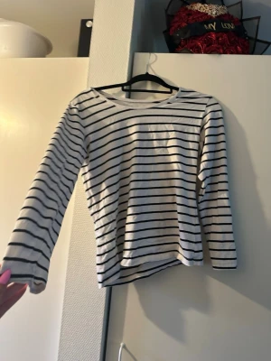Randig långärmad tröja i bomull - Säljer en vit långärmad tröja med tunna svarta horisontella ränder och rund hals. Tröjan har en bröstficka och är gjord i mjuk bomull, perfekt för en avslappnad stil.