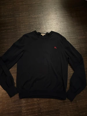 Mörkblå vintage cashmere tröja från Burberry - Klassisk mörkblå tröja från Burberry med rund halsringning och diskret röd logga broderad på bröstet. Tröjan har långa ärmar och ribbade muddar vid ärmslut och nederkant. Perfekt för en stilren och enkel look.