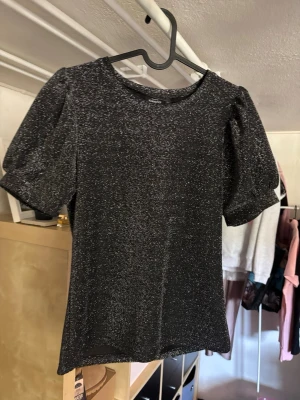 Glittrig svart topp med puffärm - Snygg svart topp med glittrigt material och puffiga korta ärmar. Perfekt för dig som vill sticka ut lite extra. Toppen har rund halsringning och sitter figurnära. Passar till både jeans och kjol för en trendig look.