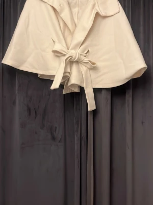 Vit cape-kappa från Gina Tricot - Stilren vit cape-kappa från Gina Tricot med bred krage och knytband i midjan. Kappan har en elegant, vid siluett och är tillverkad i ett mjukt tyg som ger en lyxig känsla. Perfekt för att lyfta din outfit under kyligare dagar.