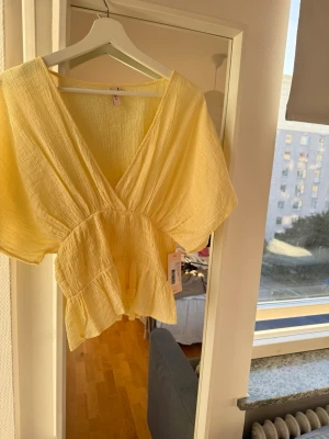 Ljusgul topp  - Supersöt ljusgul topp med v-ringning och volangdetaljer. Toppen har korta, vida ärmar och är gjord i ett lätt, strukturerat tyg som känns luftigt. Perfekt för varma dagar och ger en fräsch look.