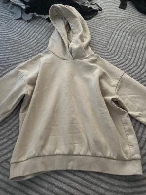 Beige oversized hoodie - En stilren beige hoodie i oversized modell med huva och breda muddar. Hoodien har en enkel design utan tryck eller logga och är tillverkad i mjukt bomullsliknande material. Perfekt för en avslappnad och trendig look.