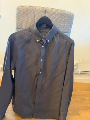 Mörkgrå skjorta från H&M - Säljer en stilren mörkgrå skjorta från H&M med regular fit. Skjortan har klassisk krage, långa ärmar och knappar framtill. Perfekt för dig som gillar enkel och clean stil.
