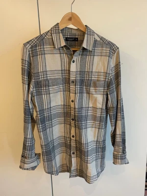 Beige och grå rutig skjorta - Säljer en beige och grå rutig skjorta med klassisk krage och långa ärmar. Skjortan har knappar framtill och en bröstficka. Perfekt för en avslappnad och stilren look.