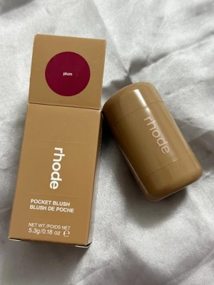 Rhode Pocket Blush Plum - Rhode Pocket Blush i färgen Plum är ett krämrouge i stickformat. Förpackningen är kompakt och beige med vit logga. Rouget har en djup plommonlila nyans som ger en fräsch och naturlig look till kinderna. Perfekt att ha med i väskan för touch-ups under dagen.