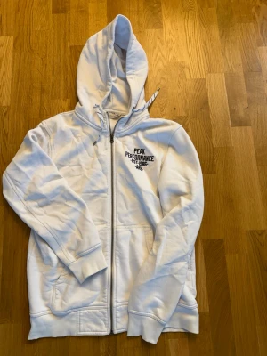Vit hoodie från Peak Performance - Säljer en vit hoodie från Peak Performance med dragkedja framtill och två fickor. Hoodien har justerbar huva med snören och svart logga på bröstet. Tillverkad i mjukt bomullsmaterial, perfekt för chill dagar eller träning.