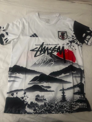 Japan landslags t-shirt med Mount Fuji - Säljer en vit sporttröja med svart och röd grafik inspirerad av japansk natur och kultur, inklusive Mount Fuji, soluppgång och tempel. Tryck på både fram- och baksida, samt Japan-emblem på bröstet. Perfekt för dig som gillar unika sportkläder med cool design.