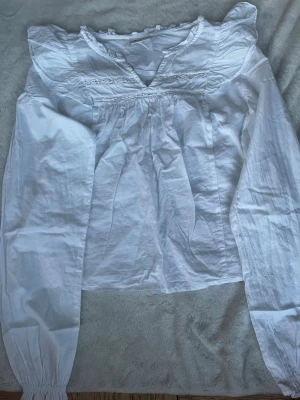 Vit romantisk blus med spetsdetaljer - Söt vit blus med långärmad design, v-ringning och fina spetsdetaljer vid halsringningen. Blusen har volangkatt och är tillverkad i ett lätt och luftigt tyg, perfekt för en trendig och avslappnad look. Har aldrig använt men saknar prislapp.