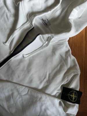 Vit zip hoodie från Stone Island - Säljer en clean vit hoodie från Stone Island med dragkedja framtill och klassisk logopatch på vänster ärm. Hoodien har huva med dragsko och ribbade muddar. Perfekt för dig som gillar streetwear och vill ha något stilrent.