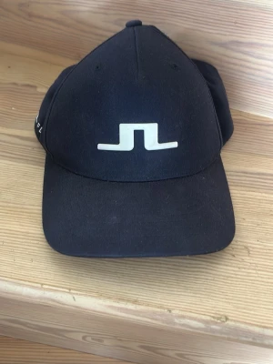 Mörkblå  keps från J.Lindeberg - Snygg mörkblå keps från J.Lindeberg med det ikoniska vita JL-logotypen framtill. Klassisk böjd skärm och justerbar baksida. Tillverkad i ett slitstarkt material och har Flexfit-teknologi för extra komfort.