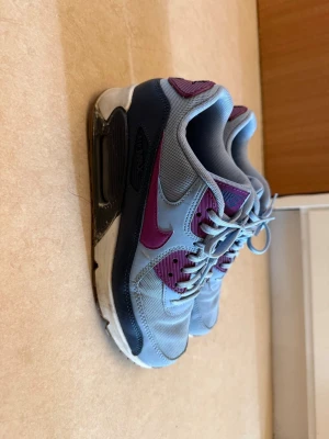 Blå och lila Nike air max sneakers - Snygga sneakers från Nike i grått med lila detaljer och swoosh. Skorna har en klassisk låg siluett, mesh och syntetmaterial på ovandelen samt en robust sula med synlig Air-enhet. Perfekta för dig som vill ha en sportig och trendig look. Bra skick men slitage syns på bilderna. Strl 40