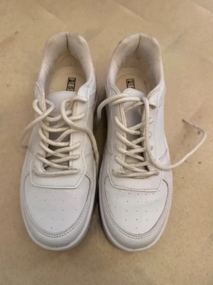 Vita klassiska sneakers - Säljer ett par vita sneakers med lågt skaft och klassisk design. Skorna har vita snören, perforerade detaljer på tån och är tillverkade i syntetmaterial. Perfekta för en clean och stilren look. Det är använda