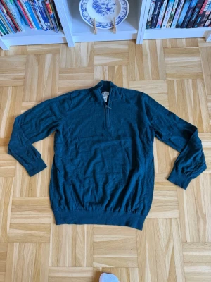 Mörkgrön tröja med halv dragkedja - Mysig mörkgrön tröja med lång ärm och halv dragkedja framtill. Tröjan har ribbade muddar vid ärmslut och nederkant samt en högre krage. Perfekt för lager på lager och enkel att matcha med jeans eller chinos.