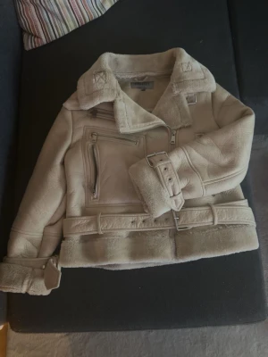 Beige teddy kappa med bälte - Snygg beige teddy kappa med bred krage, dragkedja och bälte i midjan. Jackan har detaljer som spännen vid ärmslut och axlar samt fickor med dragkedja. Perfekt för höst och vinter. Nypris 1800kr. Pris kan diskuteras 