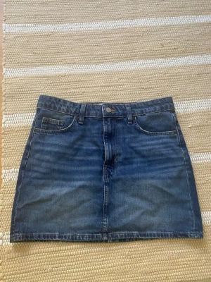 Blå jeanskjol från H&M - Säljer en klassisk blå jeanskjol från H&M i modell kortkjol. Kjolen har fem fickor, bälteshällor och stängs med knapp och dragkedja framtill. Perfekt att matcha med en enkel t-shirt eller hoodie.