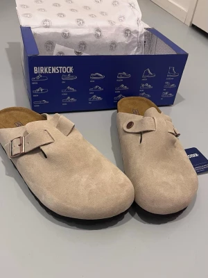 Khaki Birkenstock Bostons - Snygga beige tofflor från Birkenstock i mjuk mocka med klassisk design. De har justerbart spänne och bekväm fotbädd i kork. Perfekta för chill dagar hemma eller när du vill ha en avslappnad stil.