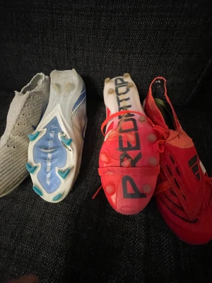 Adidas Predator och X fotbollsskor - Två par fotbollsskor från Adidas: ett par röda och vita Predator med svarta detaljer och klassisk design, samt ett par vita X med blå och turkosa detaljer. Båda har dobbar för spel på gräs och är tillverkade i syntetmaterial för bra grepp och känsla. Elit.