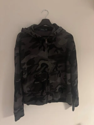 Camo hoodie - Snygg hoodie med camouflagemönster i svart och grått. Tröjan har huva, dragkedja framtill och en stor ficka på magen. Perfekt för en avslappnad streetstyle-look. Materialet känns mjukt och bekvämt.