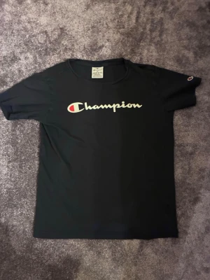 Svart Champion t-shirt med logga - Svart t-shirt från Champion med klassisk logga i vitt och rött framtill. T-shirten har rund halsringning och korta ärmar. Perfekt för en avslappnad och sportig stil.
