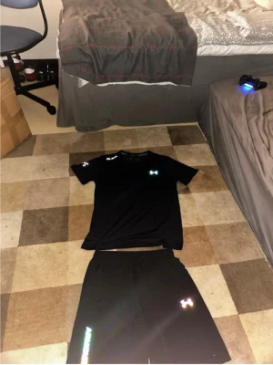 Svart träningsset från Under Armour - Säljer ett svart träningsset från Under Armour med t-shirt och shorts. Båda plaggen har reflekterande loggor och är tillverkade i ett lätt, snabbtorkande material som passar perfekt för träning eller sportaktiviteter.